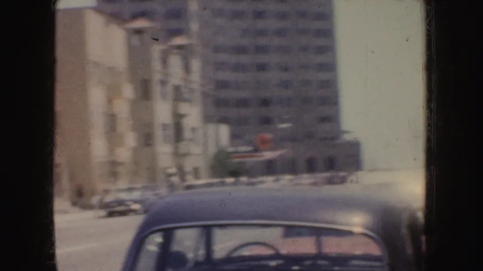 Soft 8mm view of a California downtown street with mid-century buildings, parked or moving cars, and a classic car roof in the foreground.