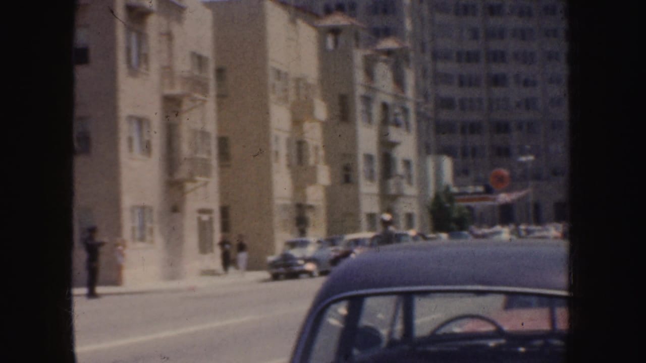 Urban street seen from a moving vehicle with parked cars, pedestrians, and mid-century buildings.