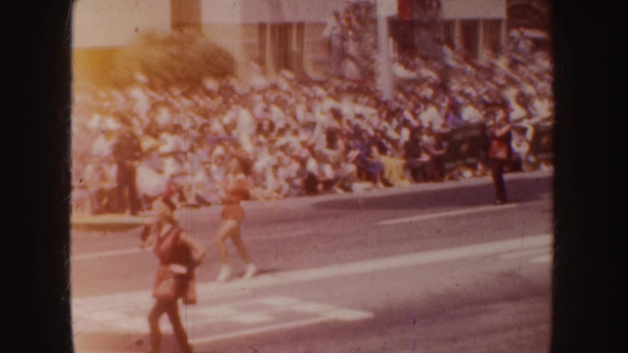 Street parade with spectators seated along the curb and performers moving through the roadway.