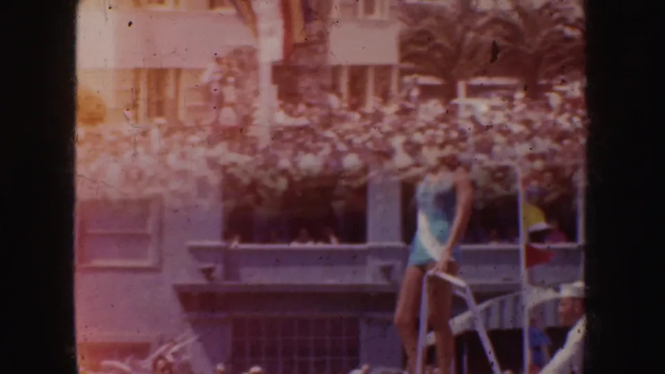 1952 California parade frame with a beauty queen on a float, crowd below, palm fronds, and urban building details in the background.