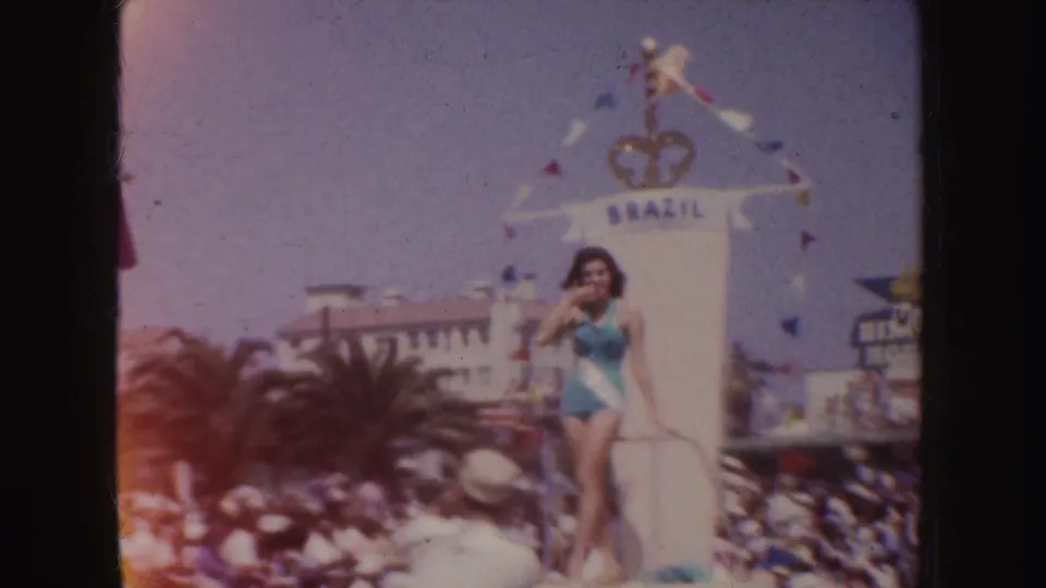 1952 California home-movie frame showing a beauty queen waving from a float marked Brazil, with palm trees and spectators visible below.
