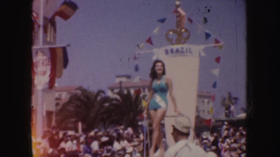 1952 California parade frame showing the beauty queen smiling and waving on a Brazil-themed float above a crowded street scene.