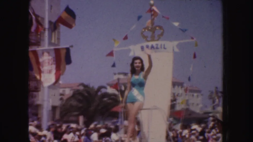 1952 California parade frame showing a beauty queen in turquoise on a float marked Brazil, with bunting, flags, and spectators below.