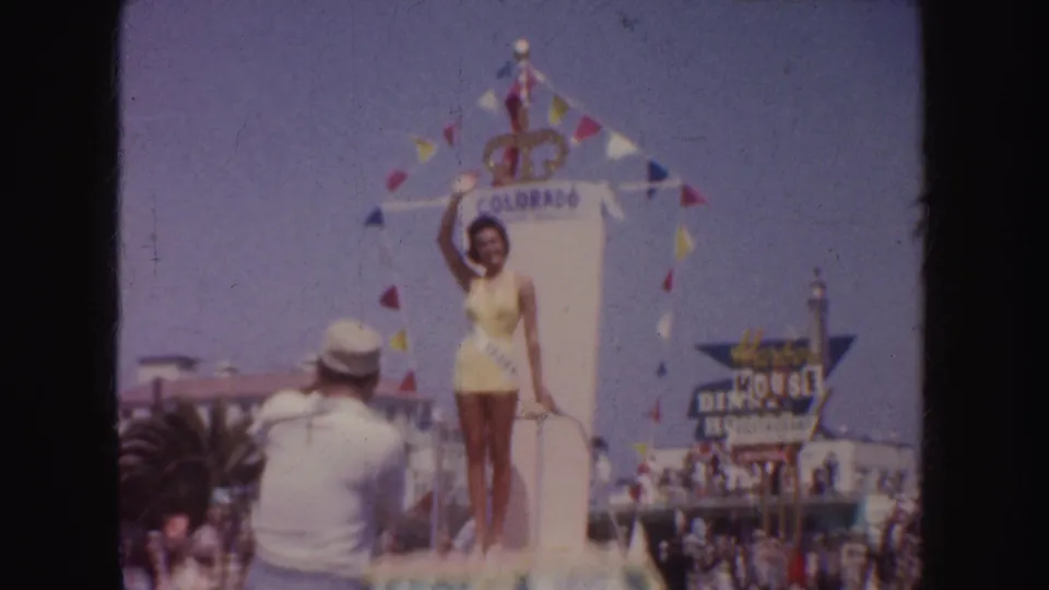 1952 California parade view of a woman in yellow on a Colorado float, with bunting, palm trees, spectators, and a nearby sign.