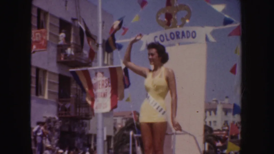 Closer 1952 California parade frame of the Colorado float queen waving, with flags, bunting, building balconies, and spectators visible.