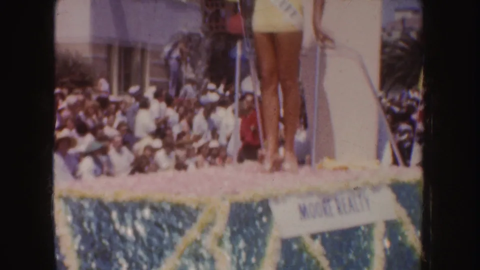 1952 California parade frame showing the Colorado float queen above a crowded street, with palm trees and decorative float trim visible.