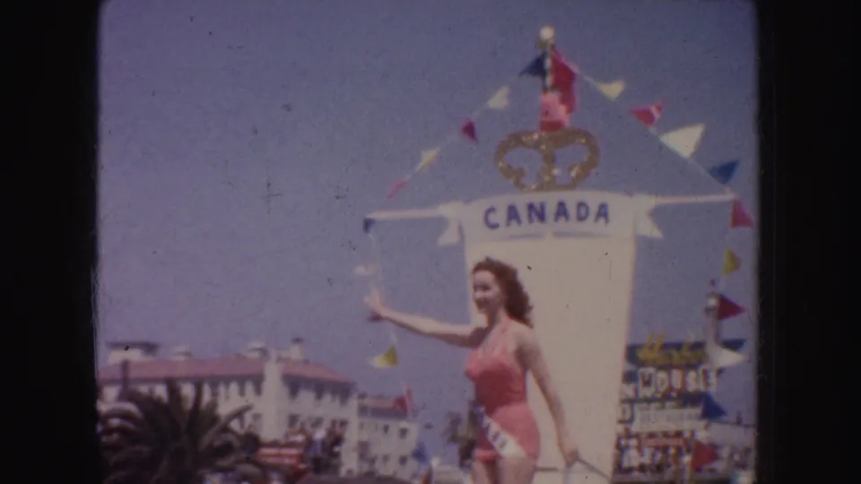 1952 California parade frame showing a woman in a pink swimsuit and sash waving beside a decorated Canada float.
