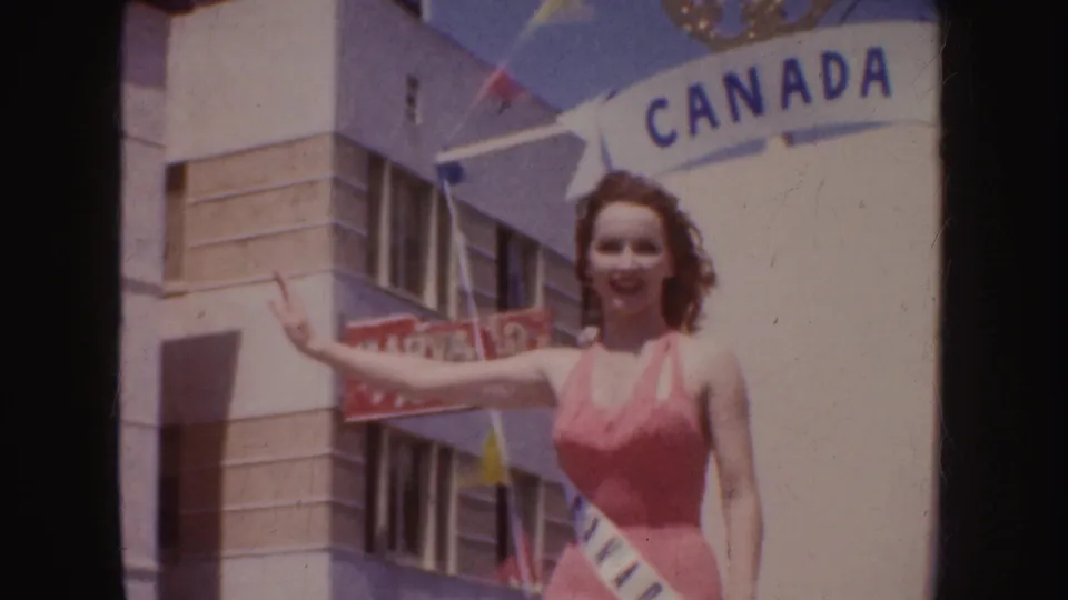 Close 1952 California parade frame with a smiling swimsuit contestant waving in front of Canada float lettering and street buildings.
