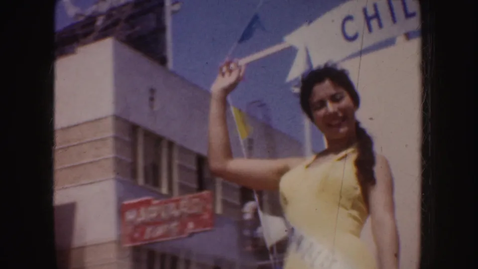 A woman in yellow swimwear and sash waves from a white CHILE parade float in sunlit 1952 California street footage.