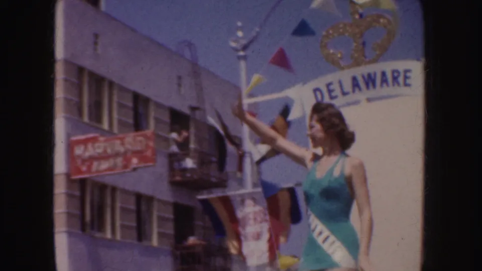 A woman in green swimwear and sash waves from a white parade float marked DELAWARE, with spectators and buildings nearby.