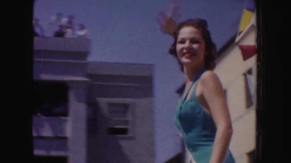 In 1952 California parade footage, a smiling woman in green swimwear and sash stands on a float under bright sky.