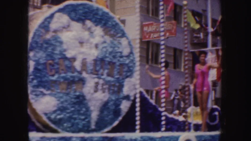 1952 California parade frame showing a float with a large globe sign reading Catalina Swim Suit and a woman in a pink swimsuit waving.