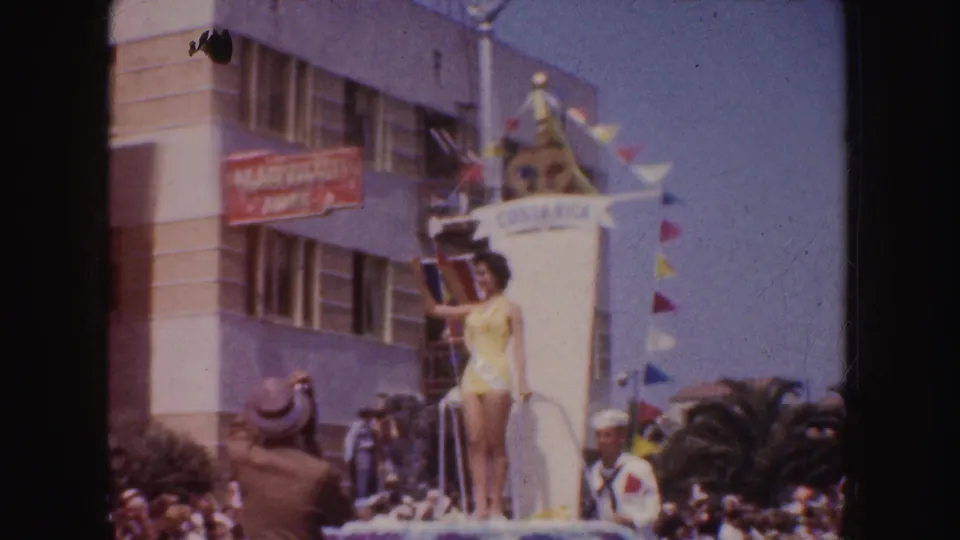 1952 California parade frame with a woman in a yellow swimsuit waving from a Costa Rica-themed float with pennant flags and spectators below.