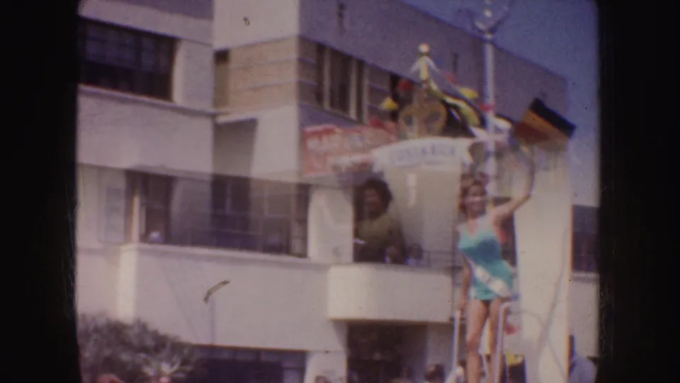 1952 California parade frame showing two women in swimsuits on a Costa Rica-themed float, with one wearing a sash near a building facade.