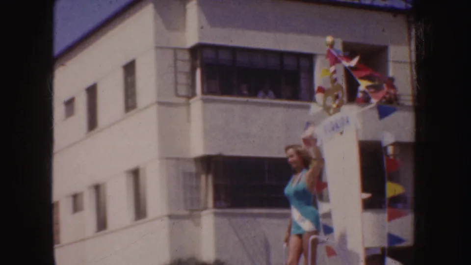 1952 California parade frame with a woman in a blue swimsuit waving beside a Costa Rica-themed float and colorful pennant flags.