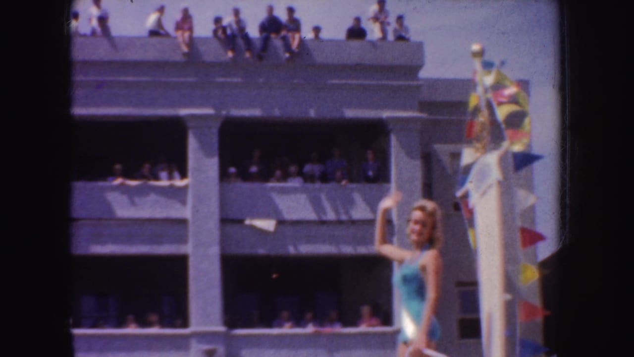 Woman in a teal swimsuit and sash waves beside a parade float while spectators watch from a building.