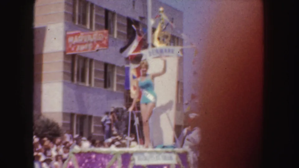 The woman stands on the Denmark-themed float, with flags, bunting, street spectators, and a readable Harvard sign in the background.
