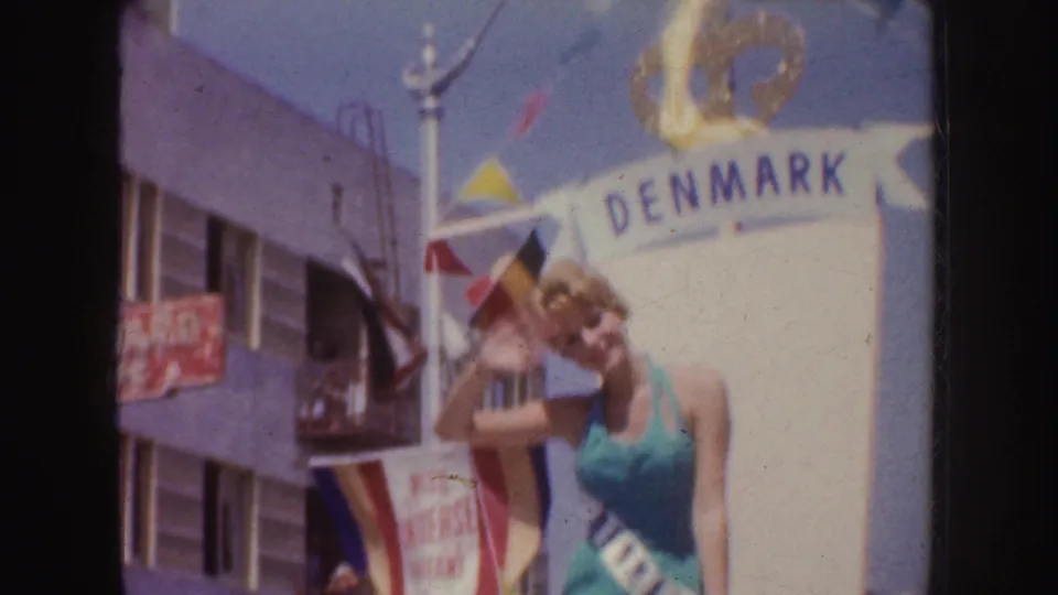 The sash-wearing woman leans forward and waves on the Denmark float, with flags and a red Harvard sign visible on the building behind.