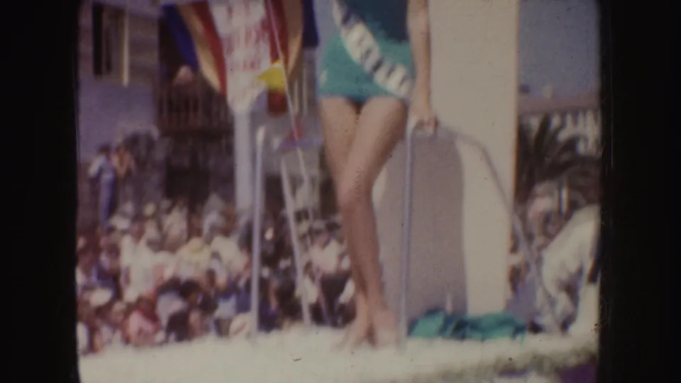 The camera frames the woman from mid-body to legs on the float platform, with the parade crowd and float decorations behind her.