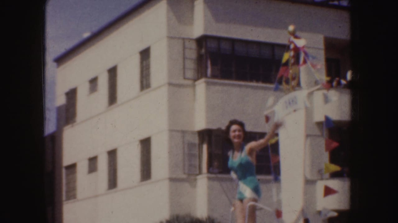 1952 California home-movie frame of a beauty queen waving from a flag-decorated parade float near a white building.