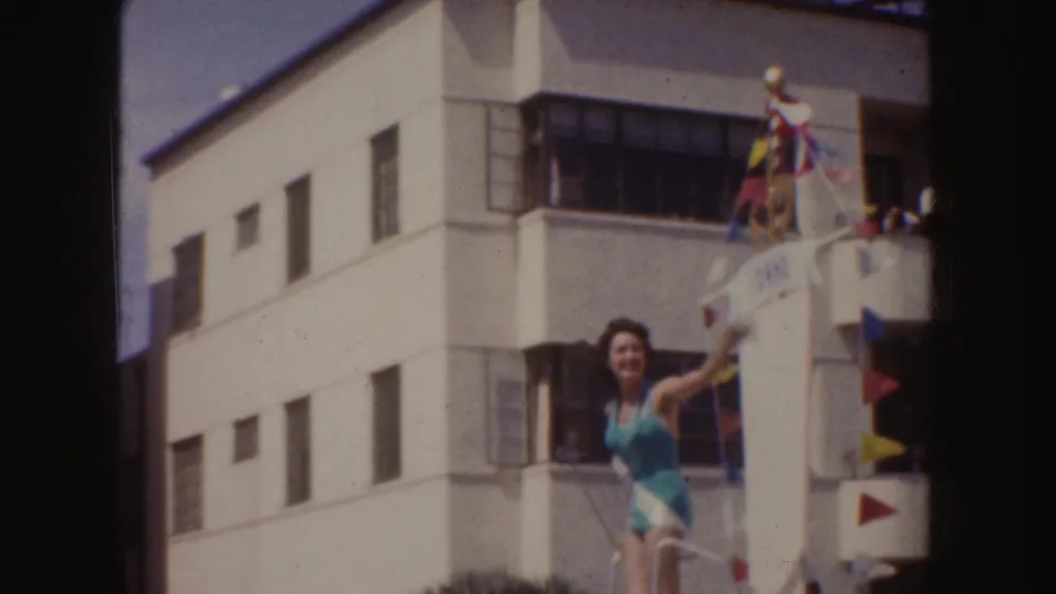 1952 California home-movie frame of a beauty queen waving from a flag-decorated parade float near a white building.