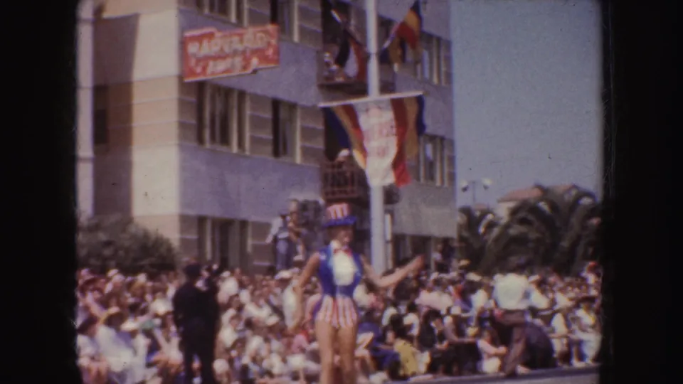 A crowd gathers along a California parade route as a flag-decorated float passes under visible streetfront signage.