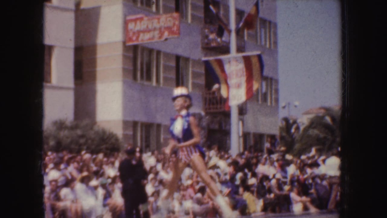 1952 California home-movie frame with a parade crowd, a woman on a float, flags, balconies, and visible street signage.