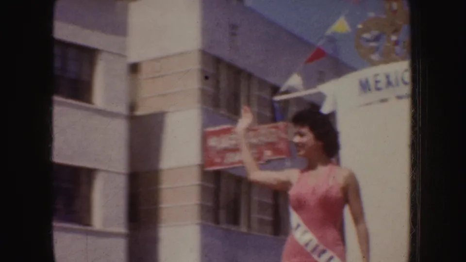 1952 California parade frame showing a sash-wearing woman in pink waving near a float marked Mexico, with buildings behind her.