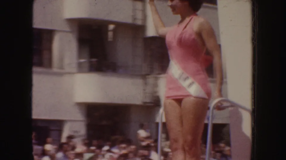 1952 California home-movie frame with a woman in a pink swimsuit and sash standing on a float before a crowd and building facade.