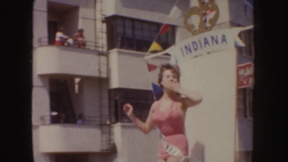 1952 California parade frame showing a sash-wearing woman near Indiana lettering, with flags, balconies, and spectators in view.