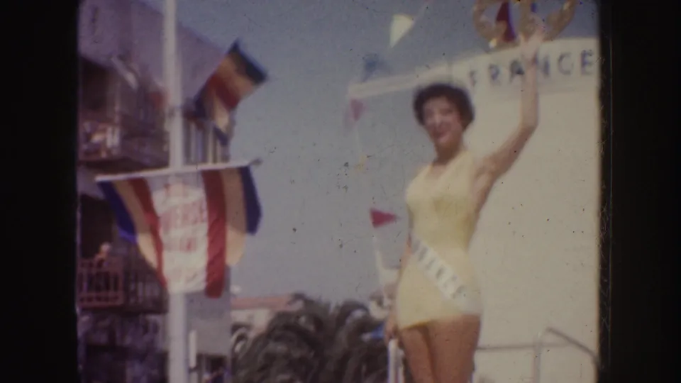 1952 California parade frame with a smiling woman in a yellow swimsuit waving beside a France sign and bunting.
