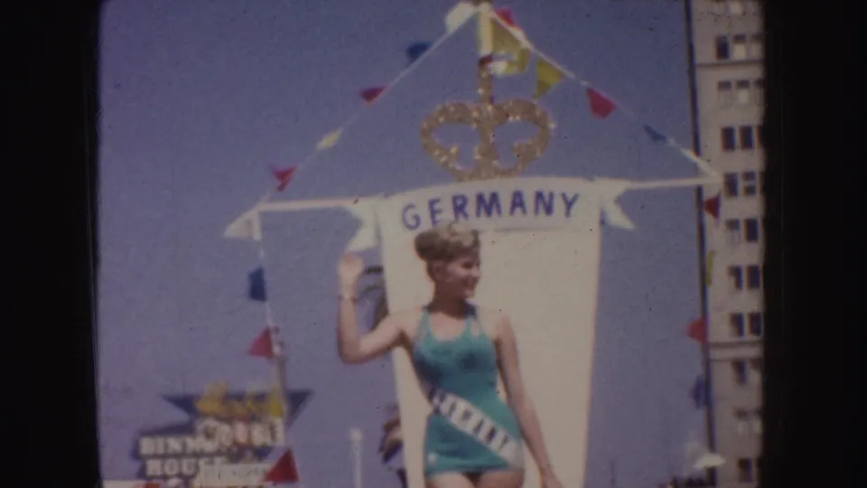 1952 California parade frame showing a woman in a teal swimsuit posing beside a Germany sign on a decorated float.