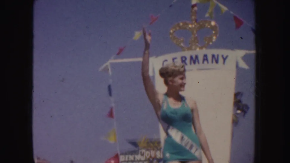 1952 California parade frame with a woman in a teal swimsuit waving on a Germany float beneath a crown emblem.