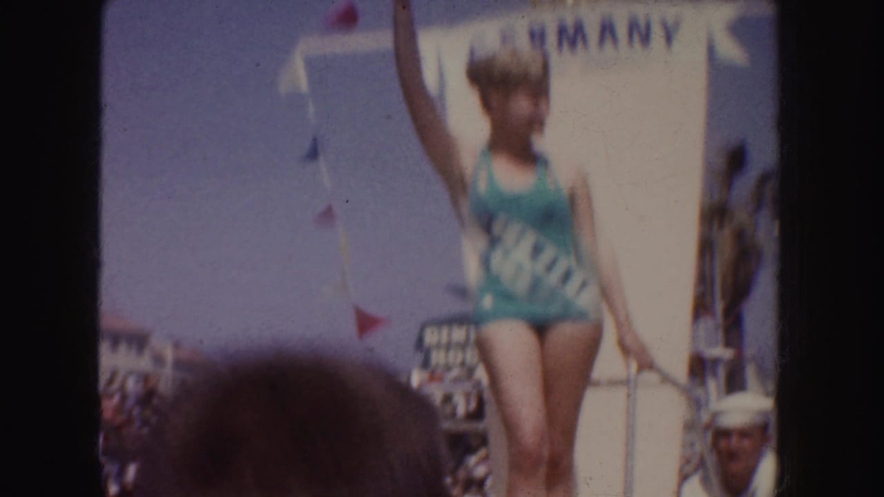 Woman in green swimsuit and GERMANY sash waves from a white parade float.