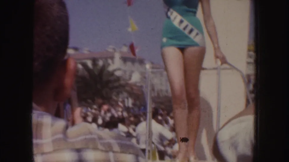 A handheld 8mm frame shows the green-swimsuit contestant partly cropped beside float railings and a nearby spectator.
