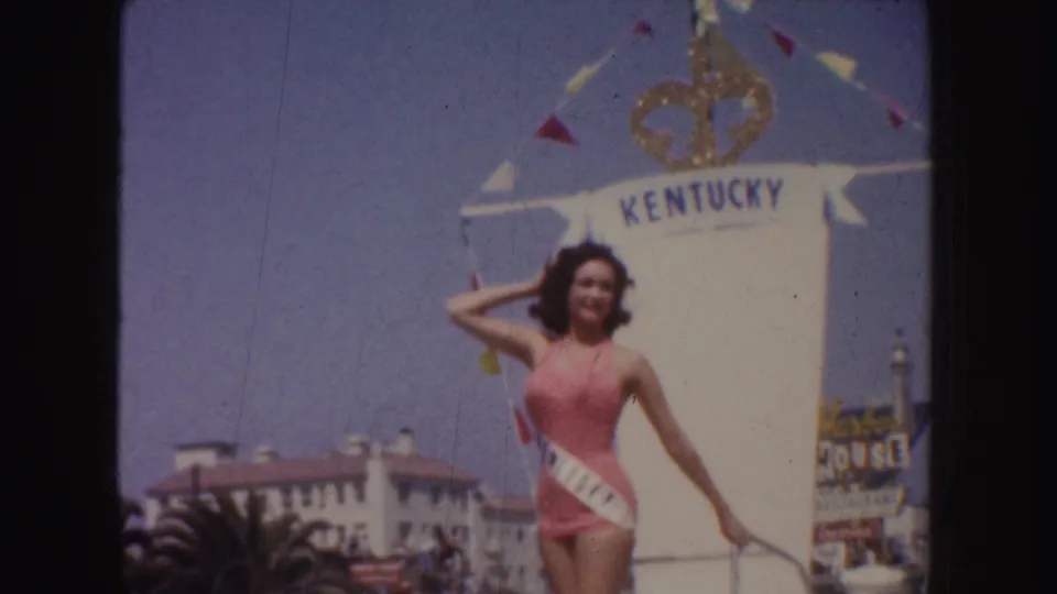 A woman in pink swimwear with a KENTUCKY sash stands near a white crowned float during the California parade footage.
