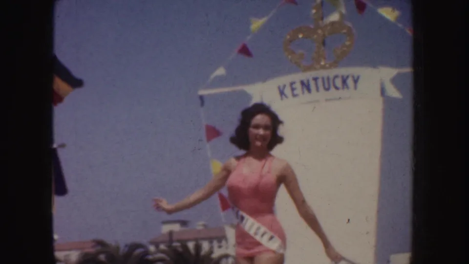 The KENTUCKY sash contestant moves past the crown-topped white float, with palm trees and buildings behind her.