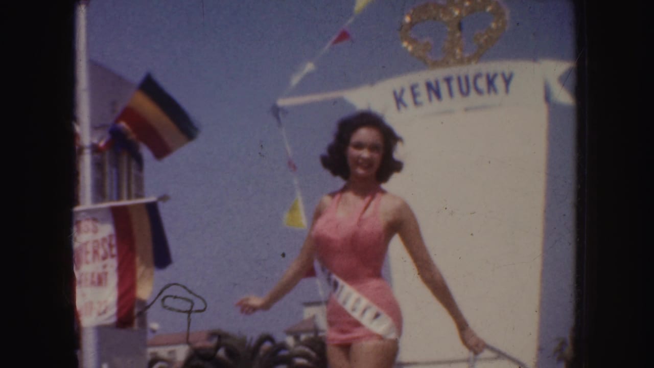 Woman in a pink swimsuit and sash standing before a Kentucky arch at an outdoor parade.