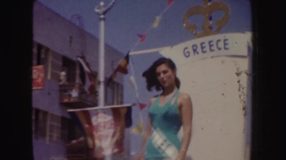 A sash-wearing woman in a teal swimsuit stands beside the GREECE arch in supported 1952 California parade footage.