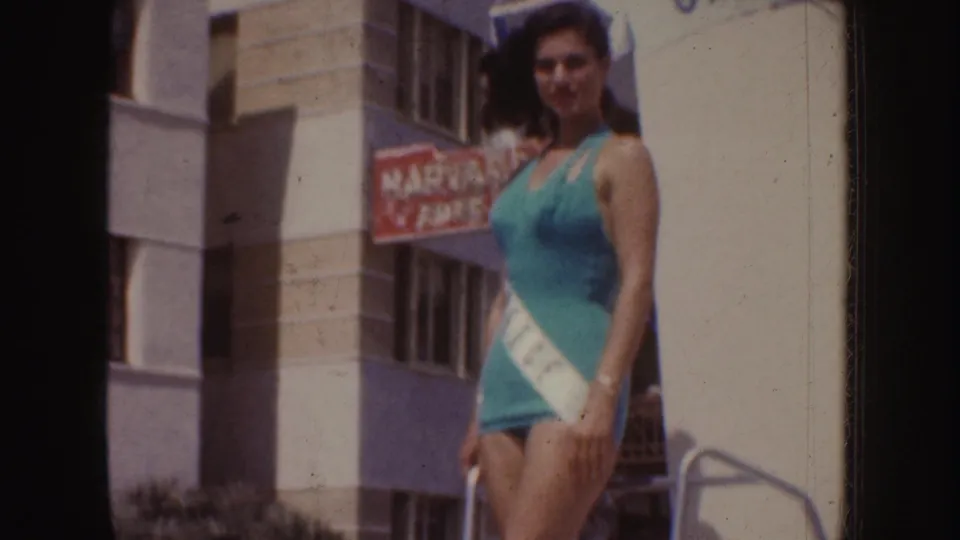 The camera moves closer to the teal-swimsuit contestant, with her sash partly visible against a mid-century building backdrop.