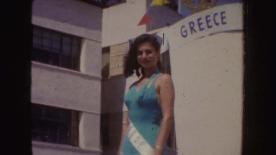 A woman in a teal swimsuit and sash smiles beside the crown-topped GREECE arch in the outdoor parade setting.