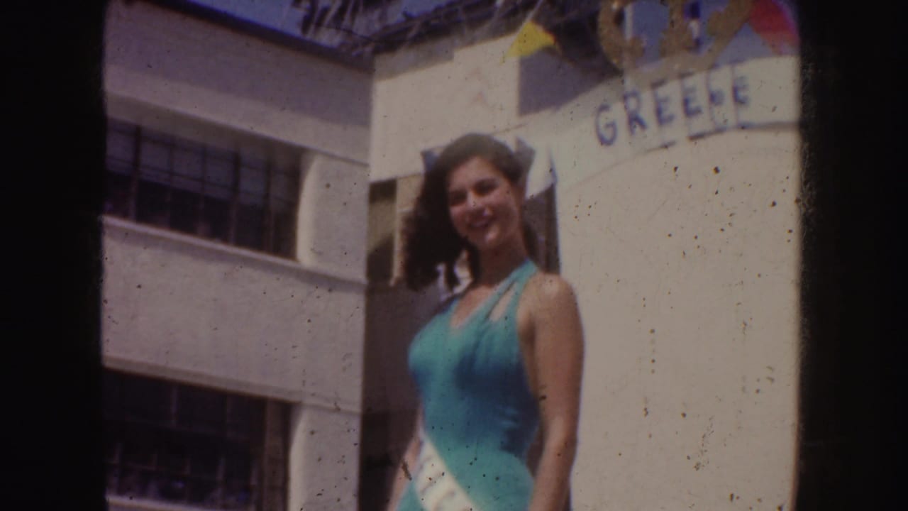 In California 1952 home-movie footage, a smiling woman in a turquoise swimsuit and sash stands near a building marked GREECE.