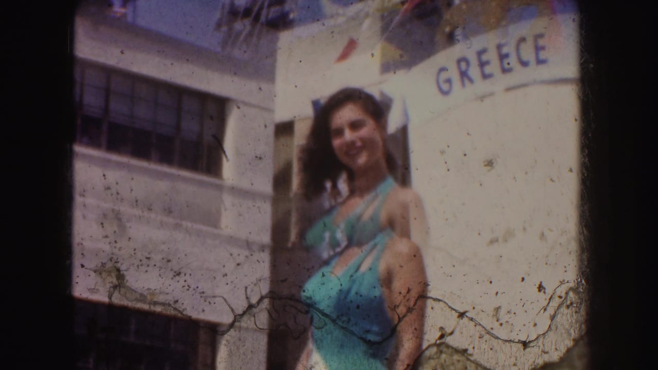 A woman smiles toward the camera beside a GREECE sign, with bunting and visible 8mm emulsion marks in the frame.