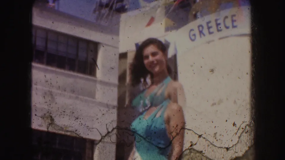 A woman smiles toward the camera beside a GREECE sign, with bunting and visible 8mm emulsion marks in the frame.