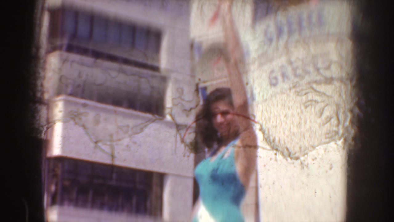 The woman raises an arm while posing near the GREECE sign in a sunlit 1952 California home-movie frame.