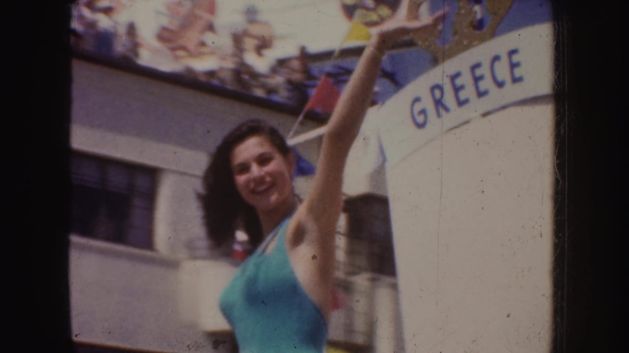 A smiling woman lifts one arm in front of a GREECE sign, with mid-century California festival decorations above her.