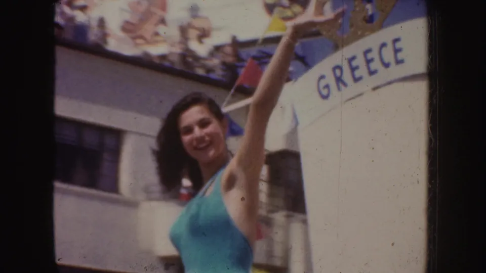 A smiling woman lifts one arm in front of a GREECE sign, with mid-century California festival decorations above her.