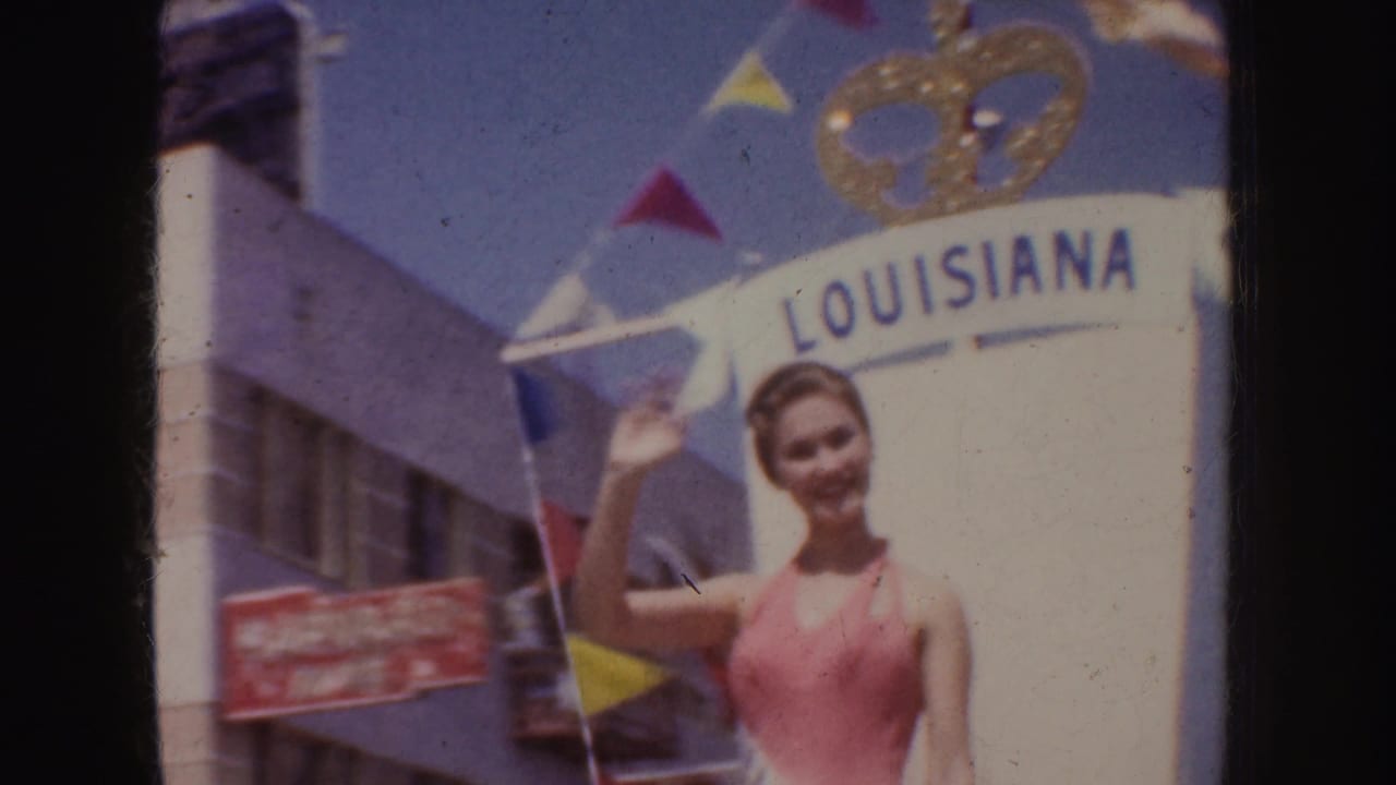Woman in a pink swimsuit waves beneath a Louisiana banner on a parade float.