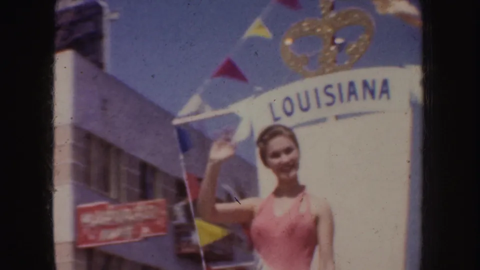 In 1952 California home-movie footage, a woman in a pink halter swimsuit and sash waves from a float marked Louisiana.