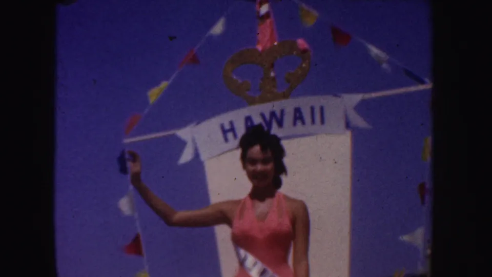 A woman wearing a pink swimsuit and sash waves from a decorated float with a Hawaii banner in 1952 California footage.